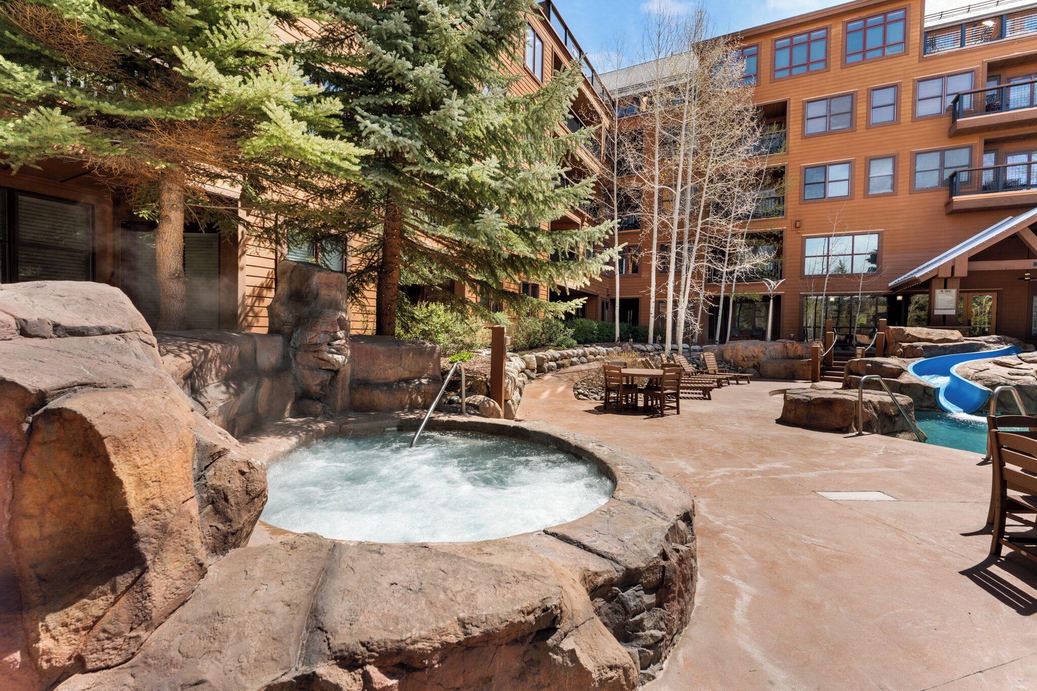 A hot tub surrounded by rocks, outdoor seating, and a pool with a slide.