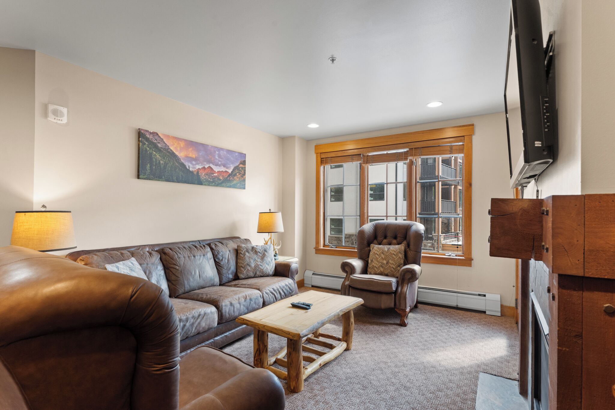 A living room with a brown leather sofa, a wooden coffee table, a leather armchair, a large window with wooden shutters, and a flat-screen TV mounted on the wall.