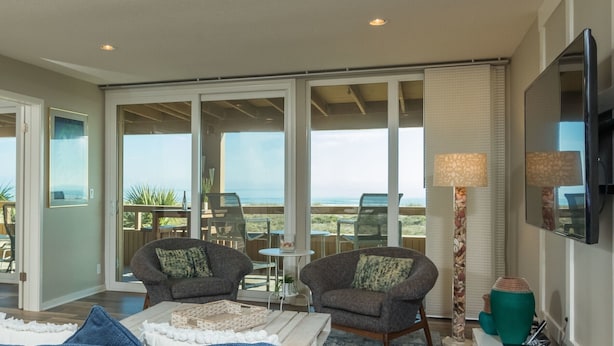 A spacious living area with two armchairs, a coffee table, and a TV mounted on the wall. Large sliding glass doors lead to a balcony with a view of the ocean. - Oceanfront - 3 Bedroom Renovated 1,800 sq ft. + Huge Balcony -  Casual Elegance (St. Augustine Beach)