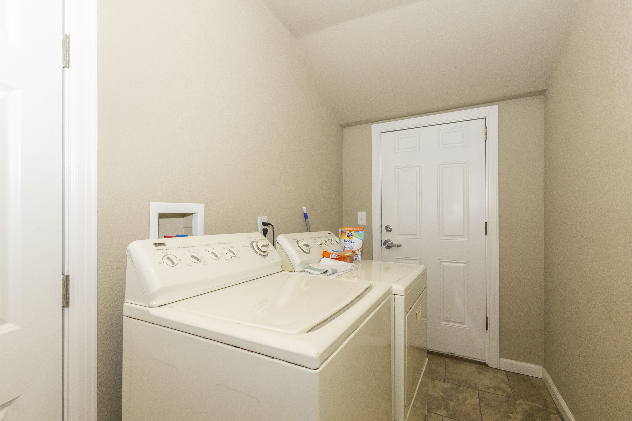 A laundry room with a front-loading washer and dryer, a door, and a small shelf with a box of detergent.
