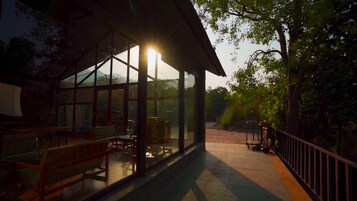 A covered outdoor seating area with tables and chairs, large windows, and a view of the surrounding trees.