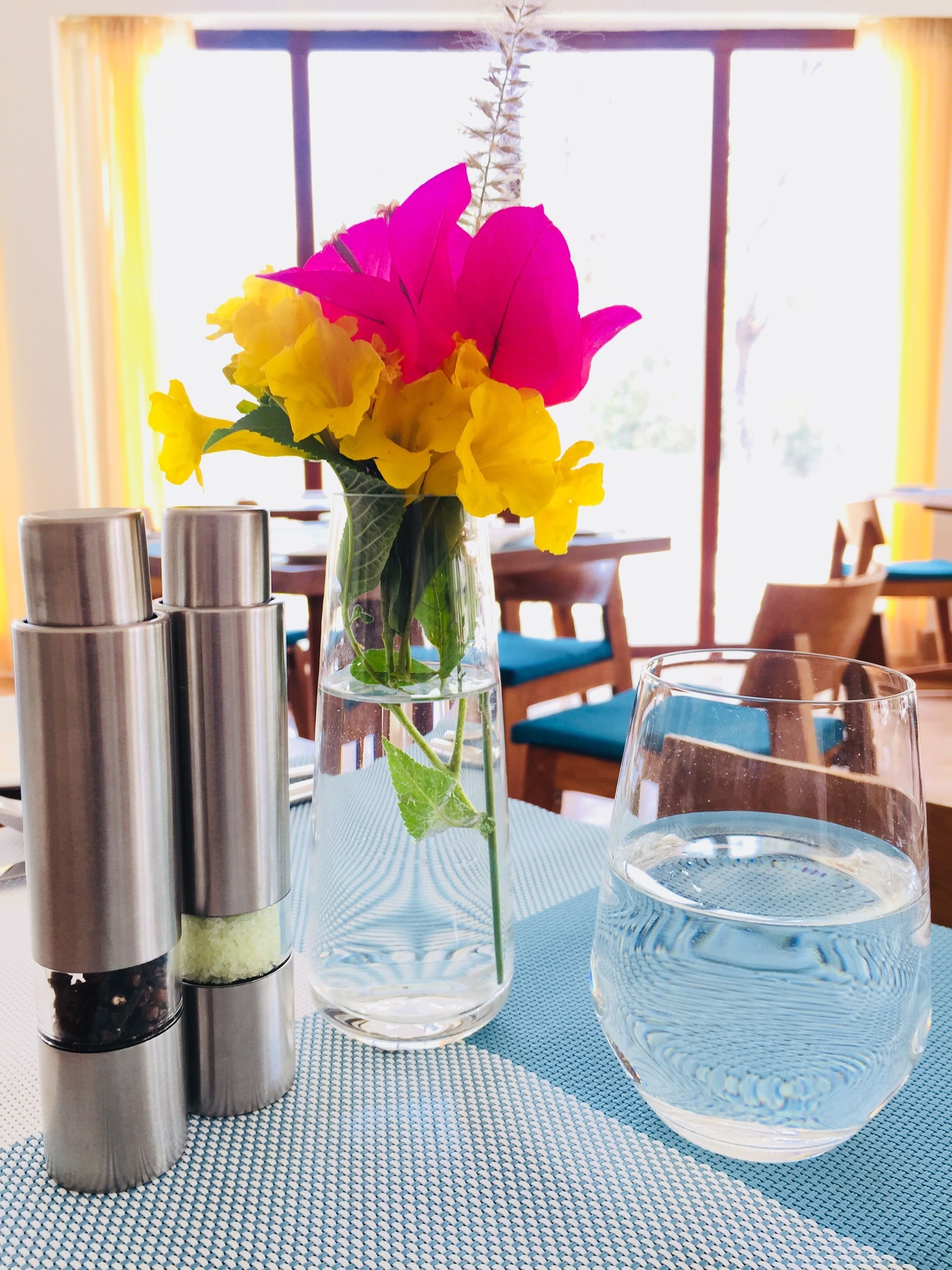 Una mesa con un florero de vidrio transparente que contiene flores amarillas y rosas, dos termos de acero inoxidable y un vaso de agua.