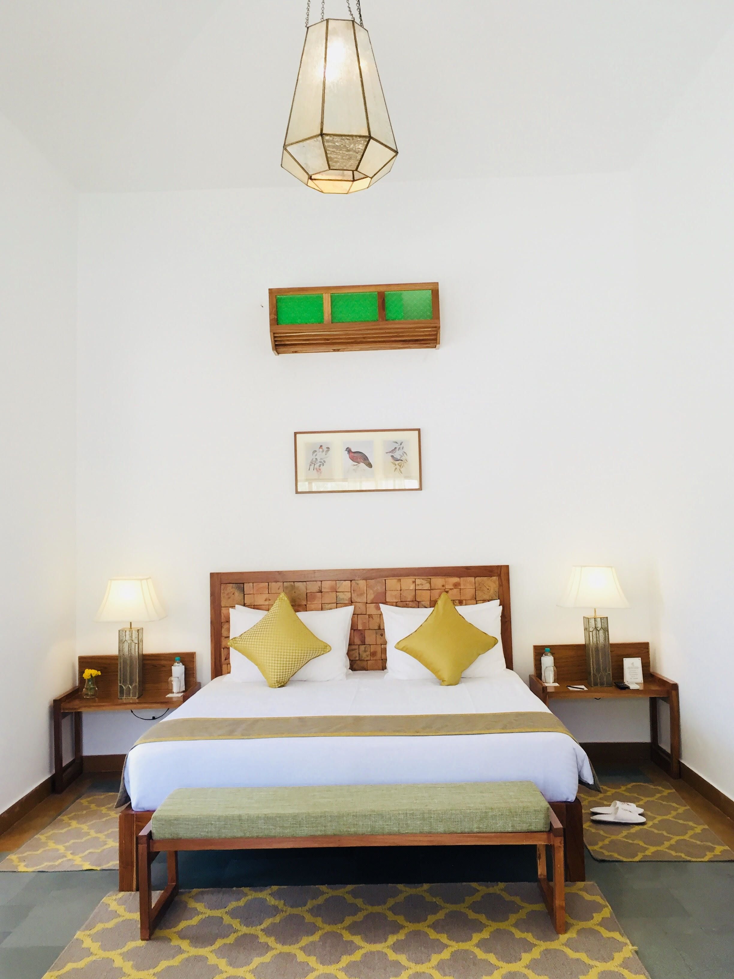 Un cuarto de hotel ordenado con una cama grande, dos mesitas de noche, una lámpara colgante, un cuadro enmarcado y un adorno verde en la pared.