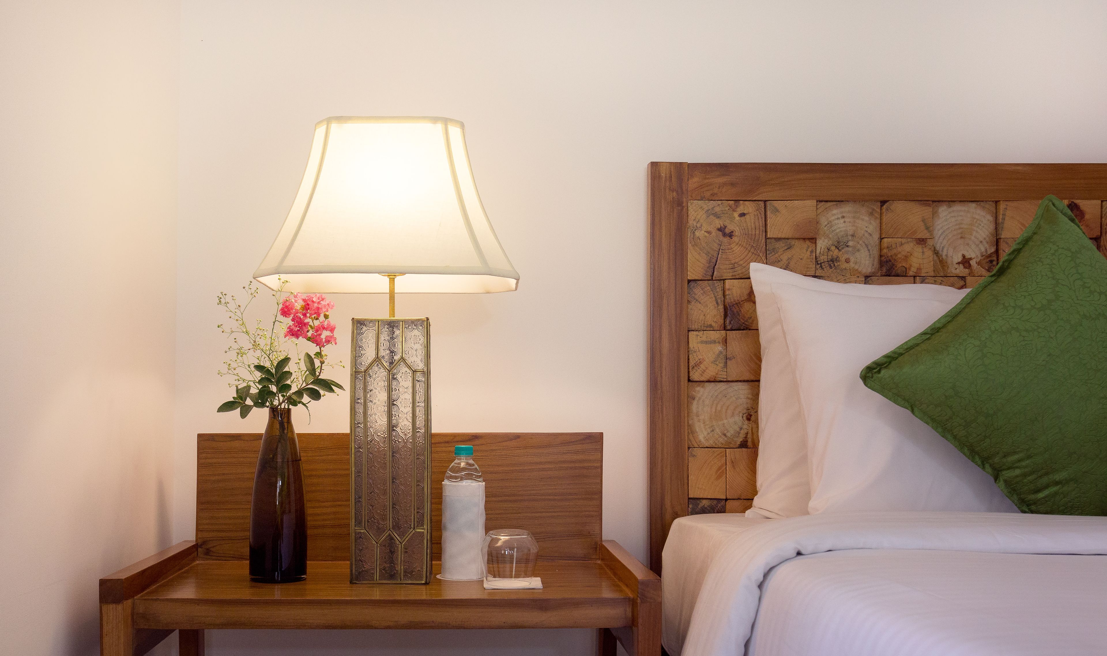 A neatly made bed with white linens and a green pillow, a wooden nightstand with a lamp, a vase with pink flowers, and a bottle of water.