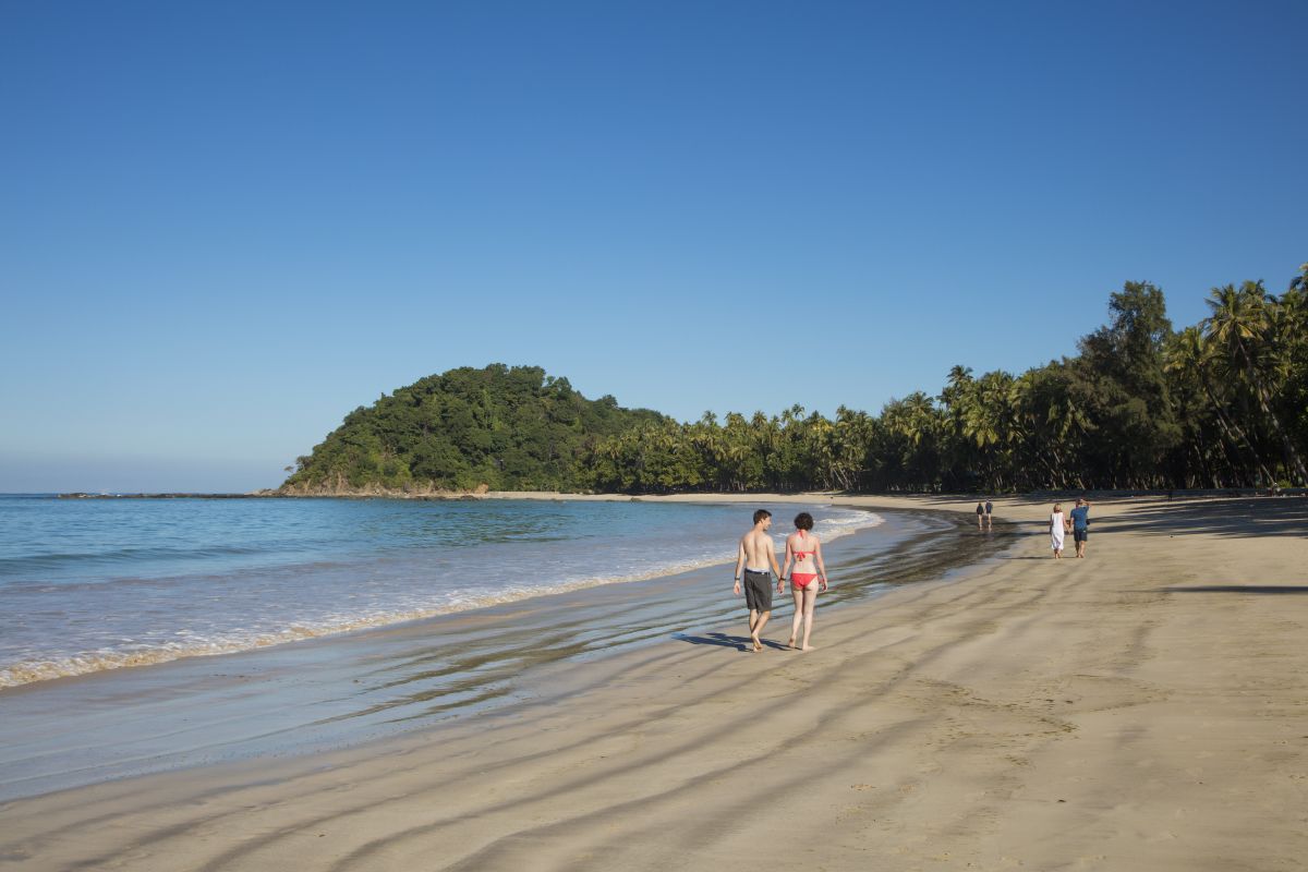 A beach with people walking along the shore, a sandy beach, and a forested hill in the background.