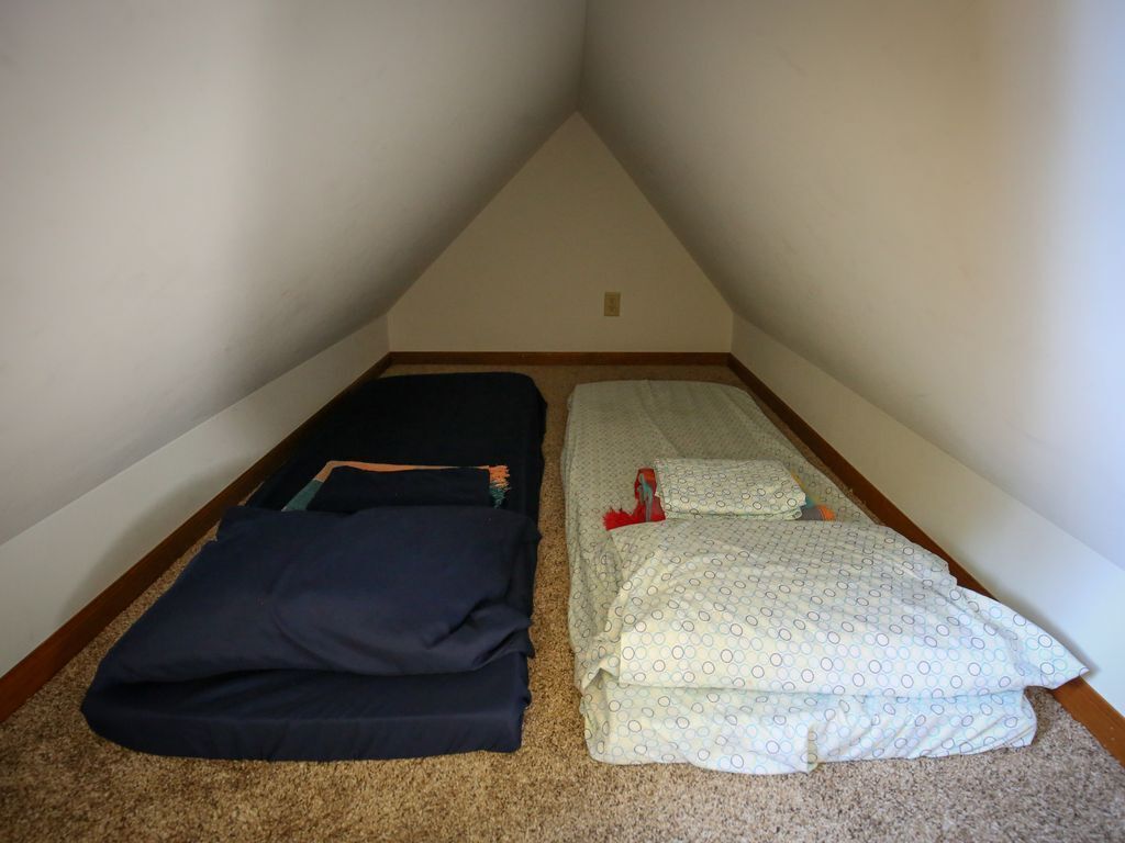 Une petite pièce mansardée triangulaire, avec deux lits : l’un est recouvert d’une couvre-lit bleu foncé, l’autre d’une couvre-lit claire à motifs.