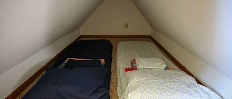Une petite pièce mansardée triangulaire, avec deux lits : l’un est recouvert d’une couvre-lit bleu foncé, l’autre d’une couvre-lit claire à motifs.