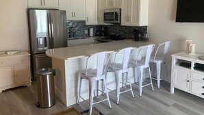 A modern kitchen with white cabinetry, a stainless steel refrigerator, a microwave, and a large island with bar stools. - Remodeled 2020  Gulf front Private condo (Santa Rosa Beach)