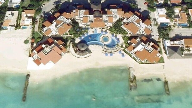 Eine Vogelperspektive zeigt einen Urlaubsort mit einem zentralen, kreisförmigen Pool, umgeben von Gebäuden, einem Strand und klarem, türkisblauem Wasser.