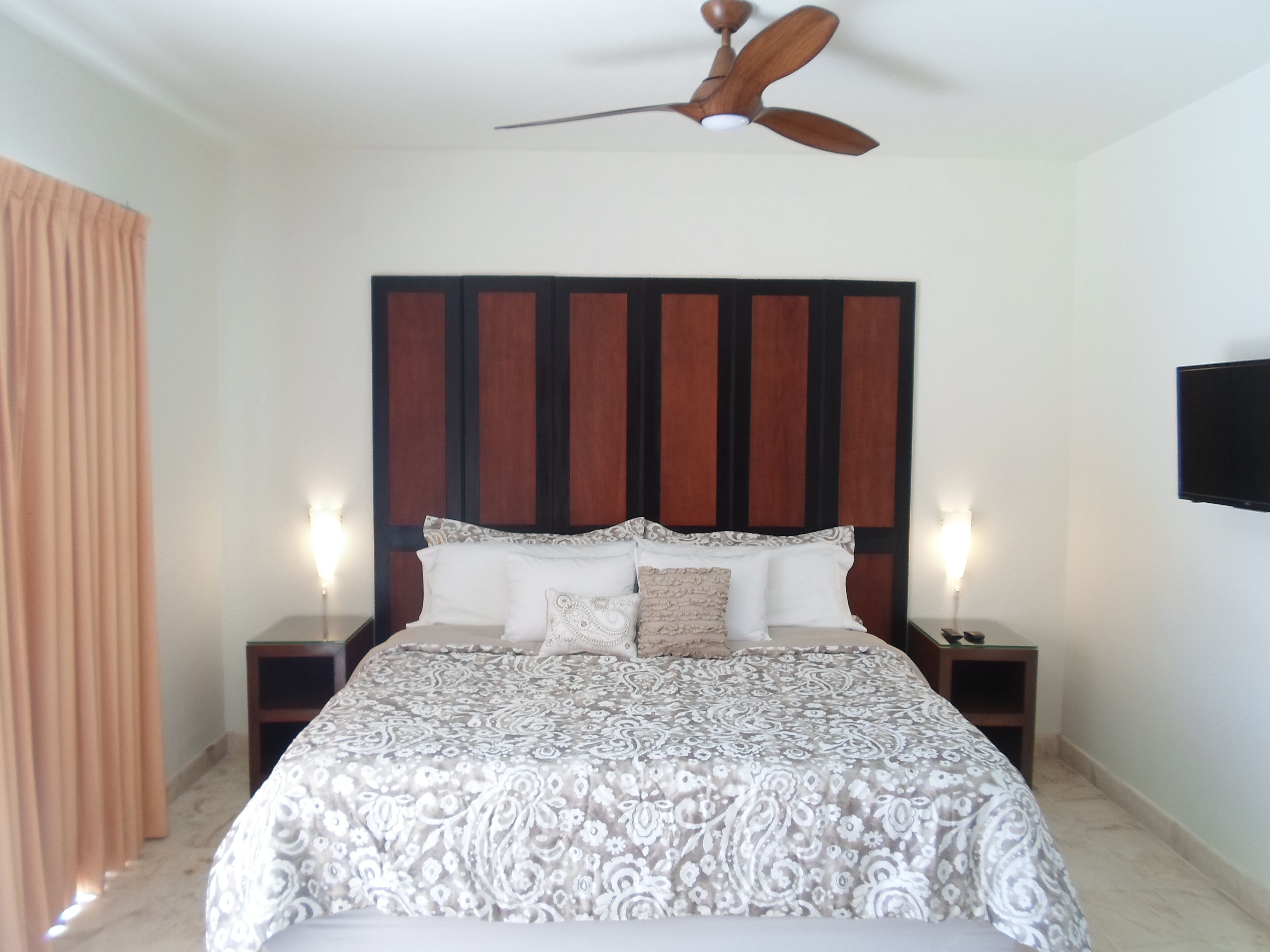 A bedroom with a large bed, a ceiling fan, and a TV mounted on the wall.