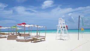 Ein Strand mit weißen Liegestühlen und Sonnenschirmen, einem Rettungsschwimmerstand und einem Volleyballnetz.