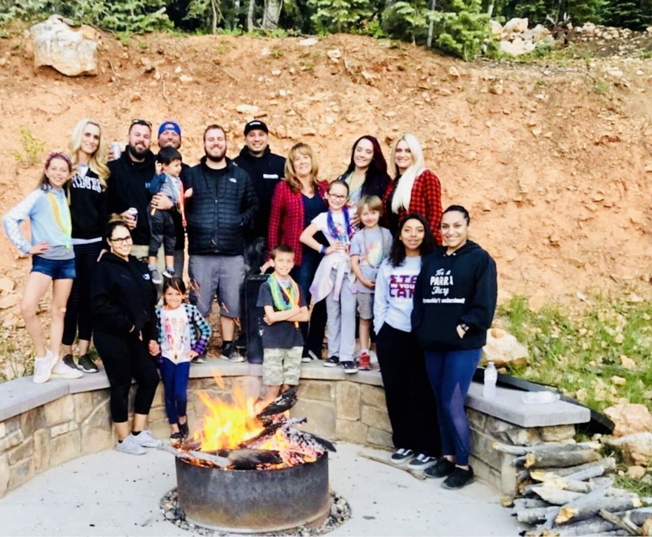 A group of people gathered around a campfire in an outdoor setting with a rocky hillside in the background.