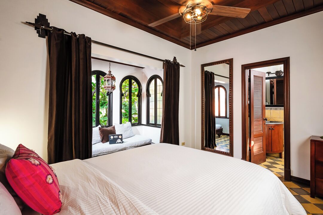 A bedroom with a large bed, dark curtains, a view of greenery outside, and a wooden ceiling fan.