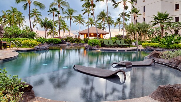 A resort pool with lounge chairs, palm trees, and a building with a thatched roof. - Relaxing 4th floor with Lap Pool View - Ocean Tower Beach Villa (Kapolei)
