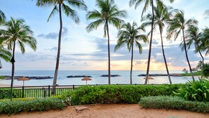 A beach view with palm trees, a fence, and a thatched umbrella. - Relaxing 4th floor with Lap Pool View - Ocean Tower Beach Villa (Kapolei)