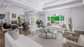 A spacious living room with a white sofa, a coffee table, and a piano. There are large windows with a view of greenery outside.