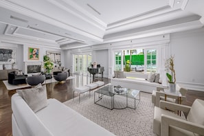 A spacious living room with a white sofa, a coffee table, and a piano. There are large windows with a view of greenery outside. - Family Vacation Dream: Privately Gated Mansion Tennis Court and Walk to Rodeo Dr (Beverly Hills)