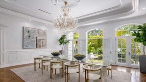 Een eetkamer met een glazen tafel, witte stoelen, een kroonluchter en grote ramen met uitzicht op groen.