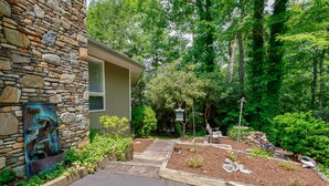 A stone building with a wooden deck and a garden with various plants and a statue. - Bella Regalo, Private 2BR, 2BA Mountain Escape, Upgraded with Panoramic Views (Pisgah Forest)