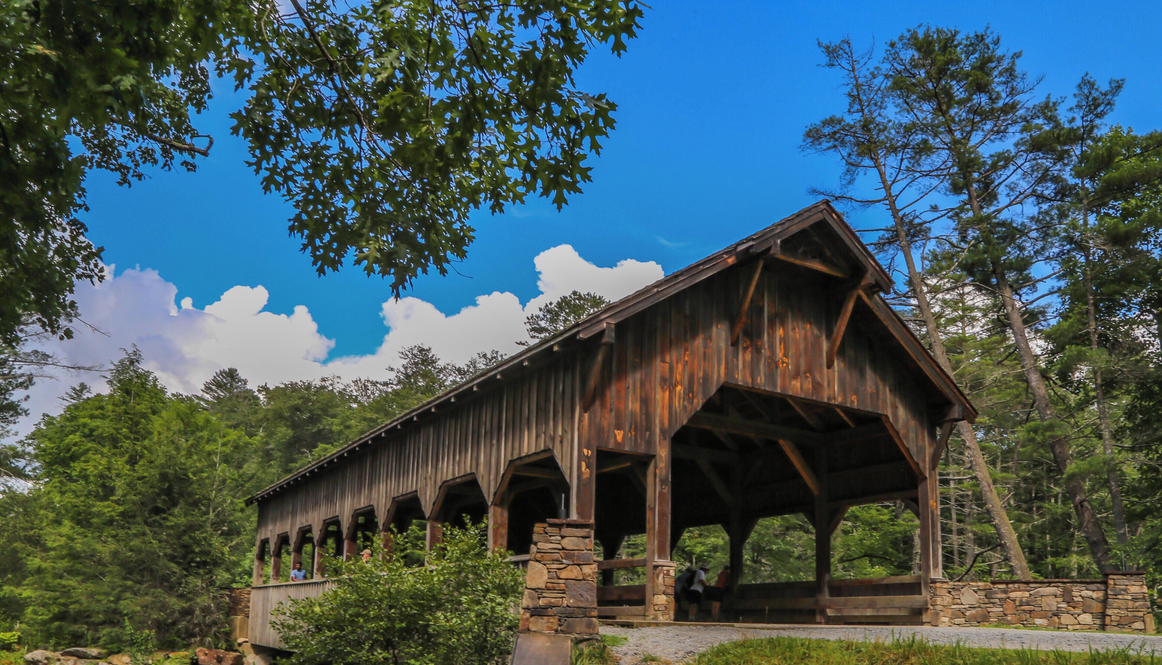 A covered wooden bridge with stone pillars, surrounded by lush greenery and a clear blue sky.