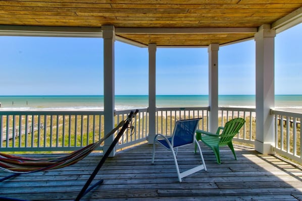 Una terraza de madera techada con vista a la playa, dos sillas y una hamaca.