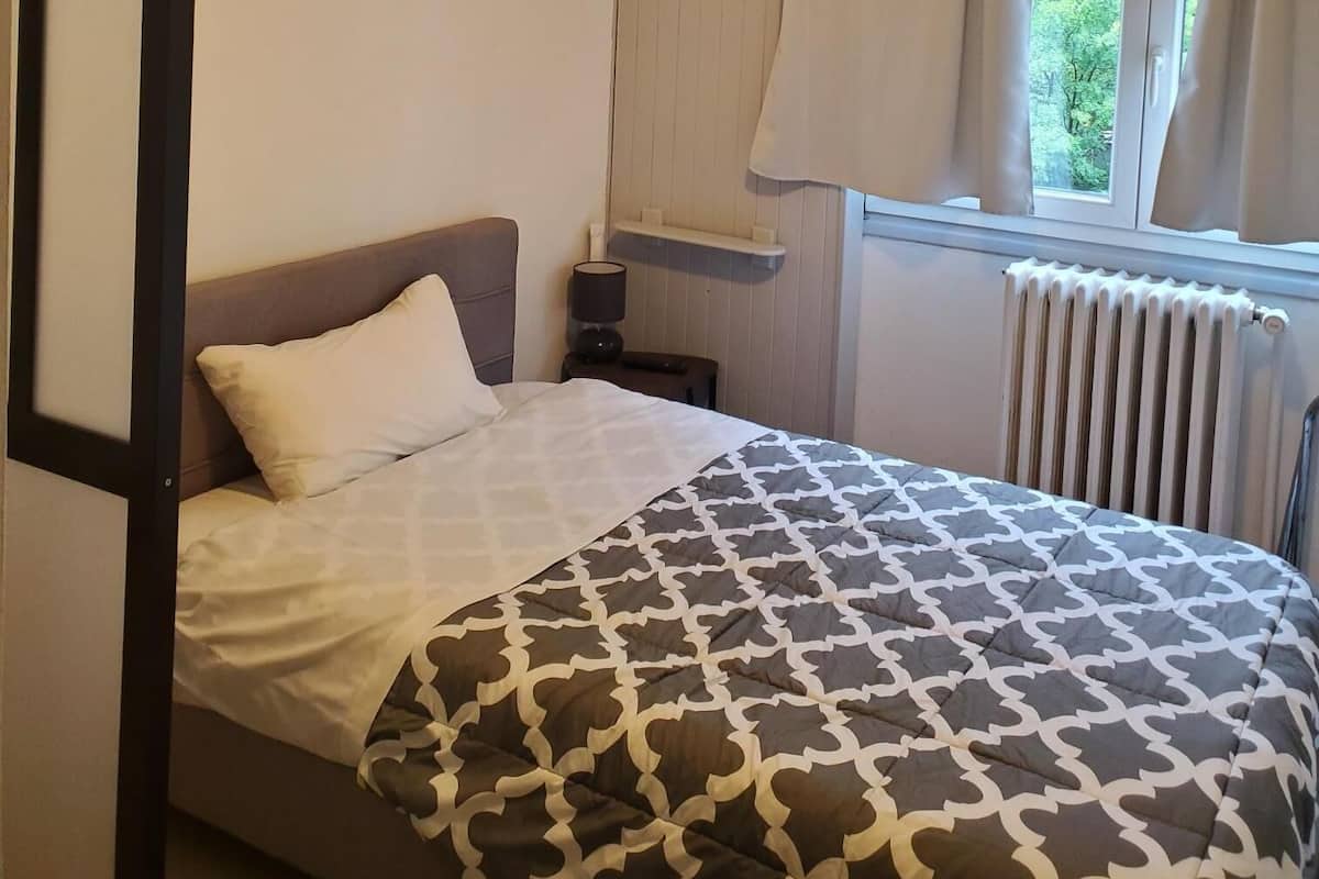Un lit simple avec une couvre-lit à motifs, une table de chevet avec une lampe, une fenêtre avec des rideaux et un radiateur.
