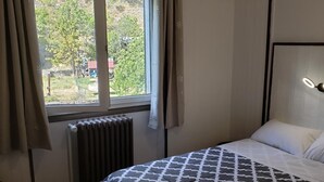 A bedroom with a bed, a window with curtains, a radiator, and a headboard with a light. - Hotel de la Gare (Modane)