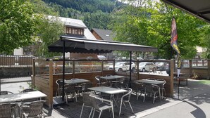 An outdoor cafe with tables and chairs under a wooden canopy, set against a backdrop of a mountain and parked cars. - Hotel de la Gare (Modane)