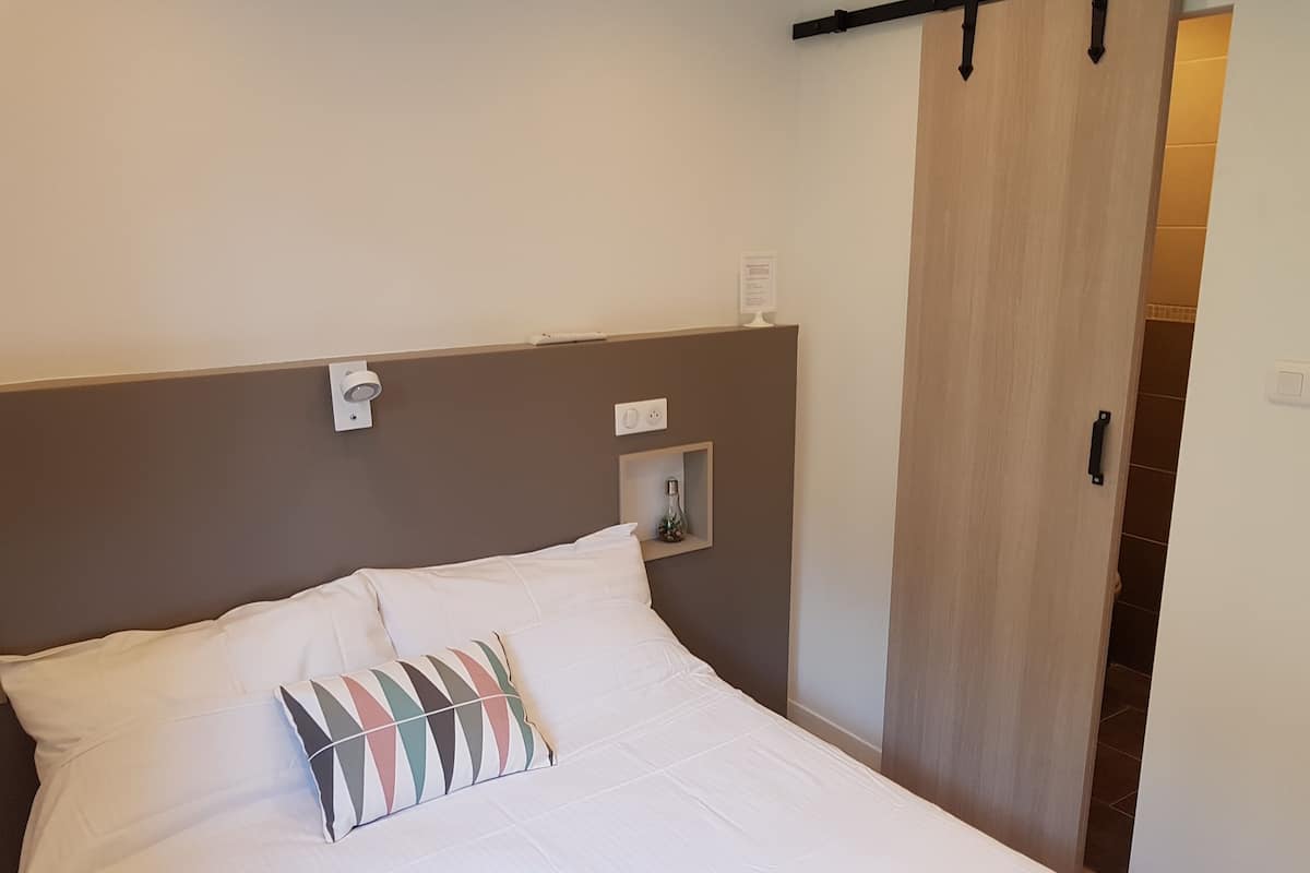 Un lit bien fait, avec du linge de lit blanc et un coussin multicolore, une petite table de chevet avec une bouteille, et une porte coulissante sur le côté droit.