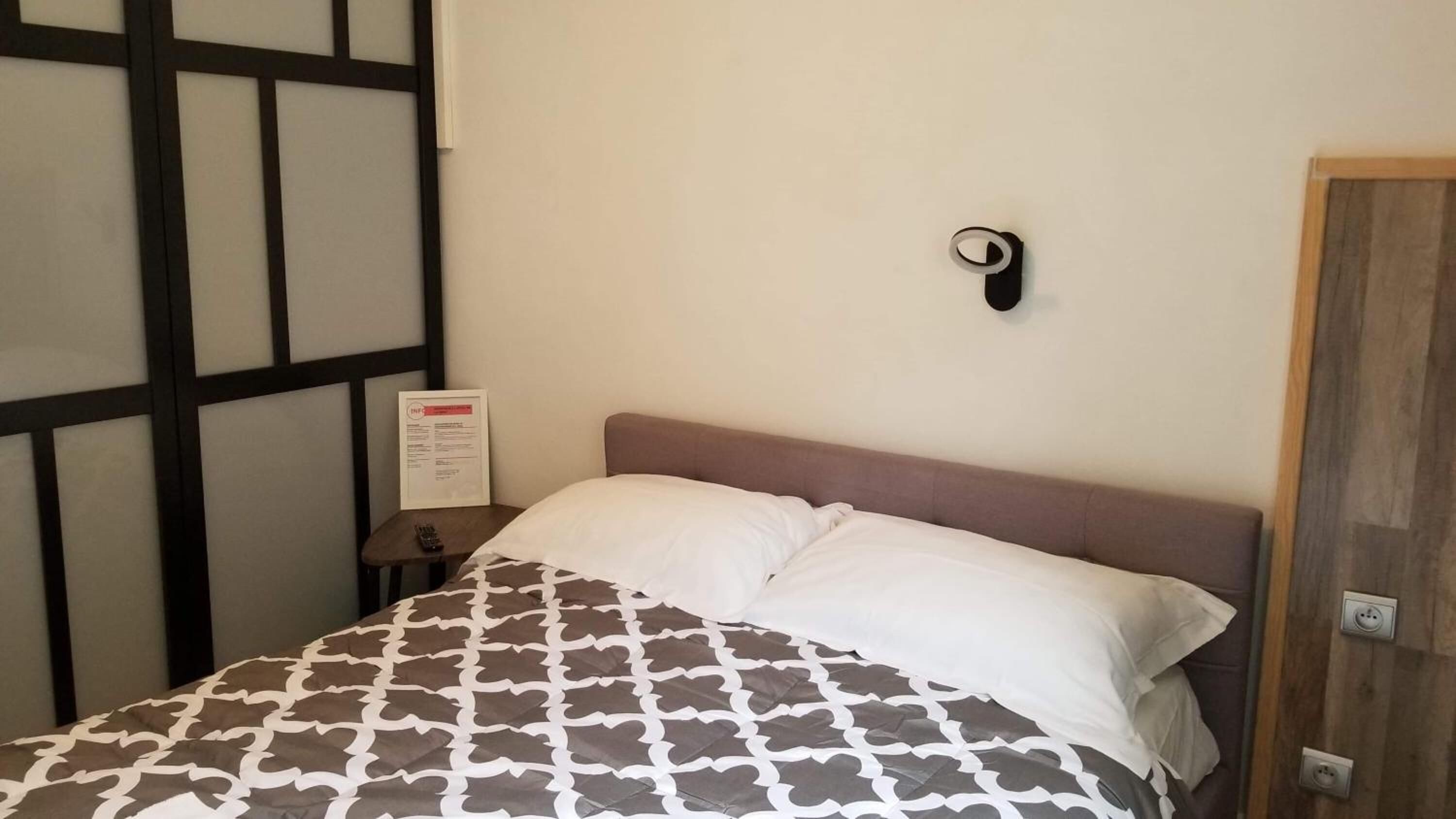 A bedroom with a bed, a nightstand, a wall-mounted light, and a door.