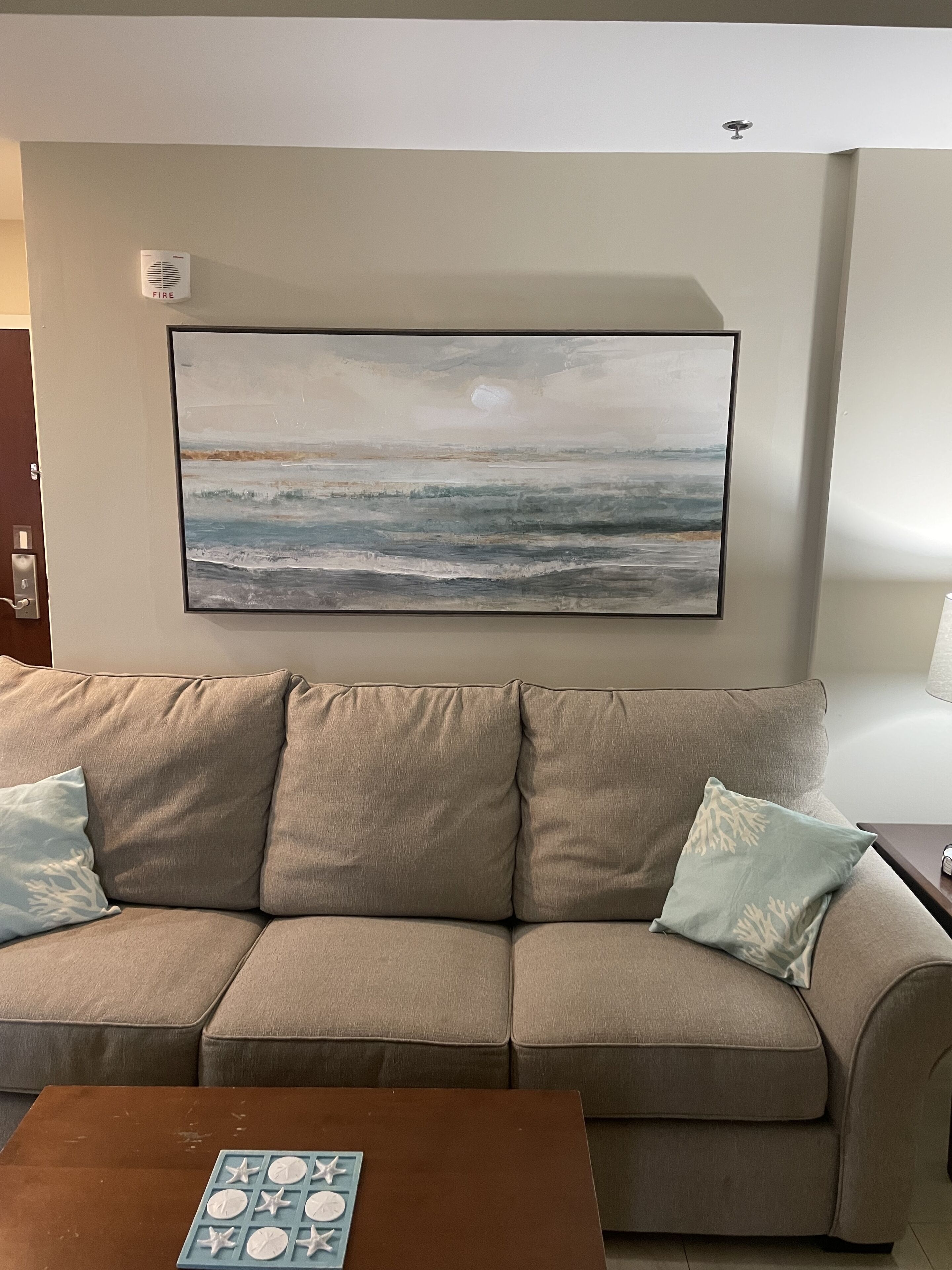 A living room with a beige sofa, two pillows, a coffee table, and a painting of a beach scene.