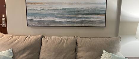 A living room with a beige sofa, two pillows, a coffee table, and a painting of a beach scene.