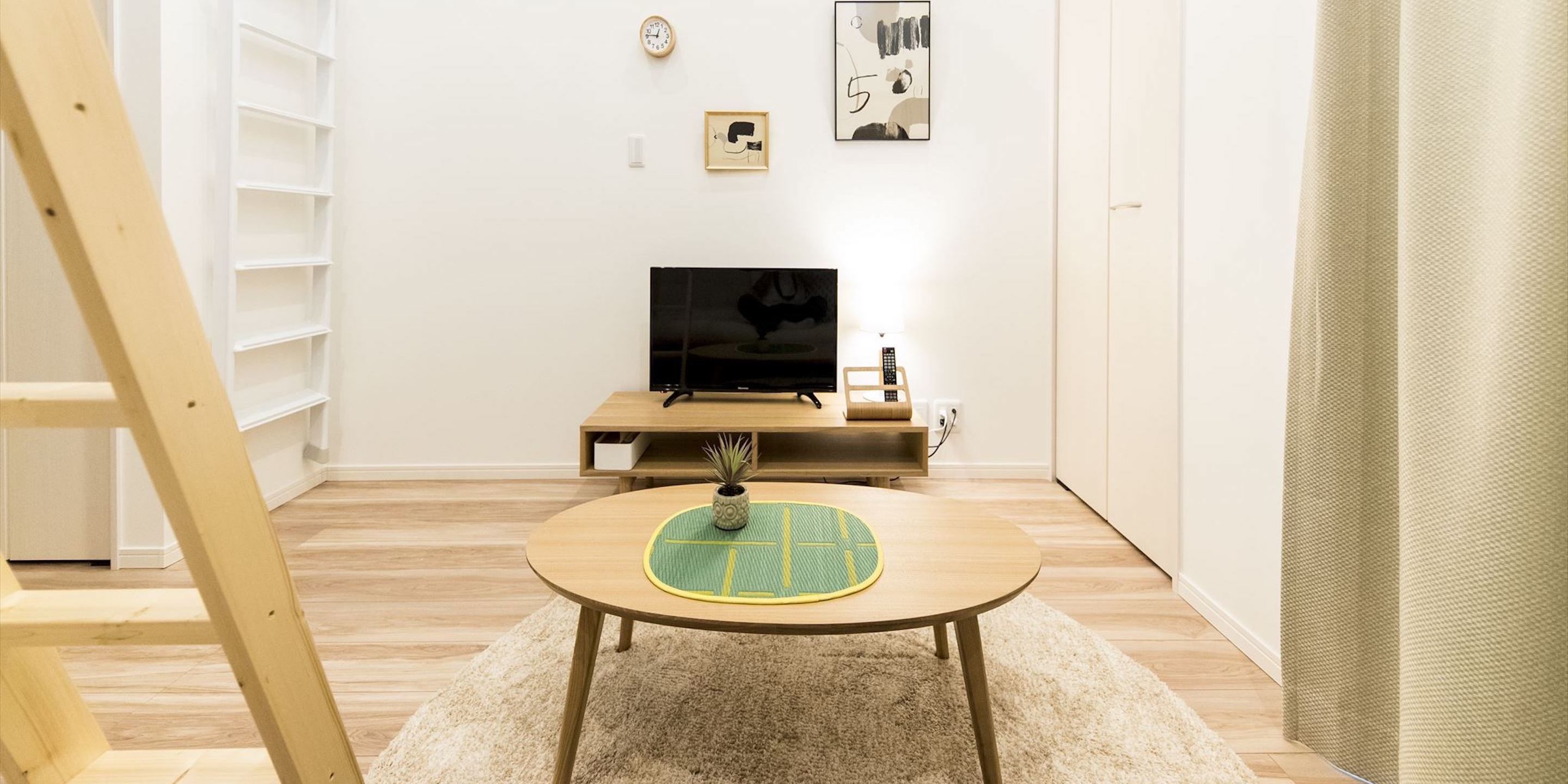 A room with a wooden floor, a round table with a green and yellow mat, a television on a wooden stand, and a ladder in the corner.
