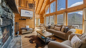 L’intérieur chaleureux d’un chalet avec une cheminée en pierre, des murs en bois, une grande fenêtre donnant sur un paysage montagneux et un coin salon confortable.