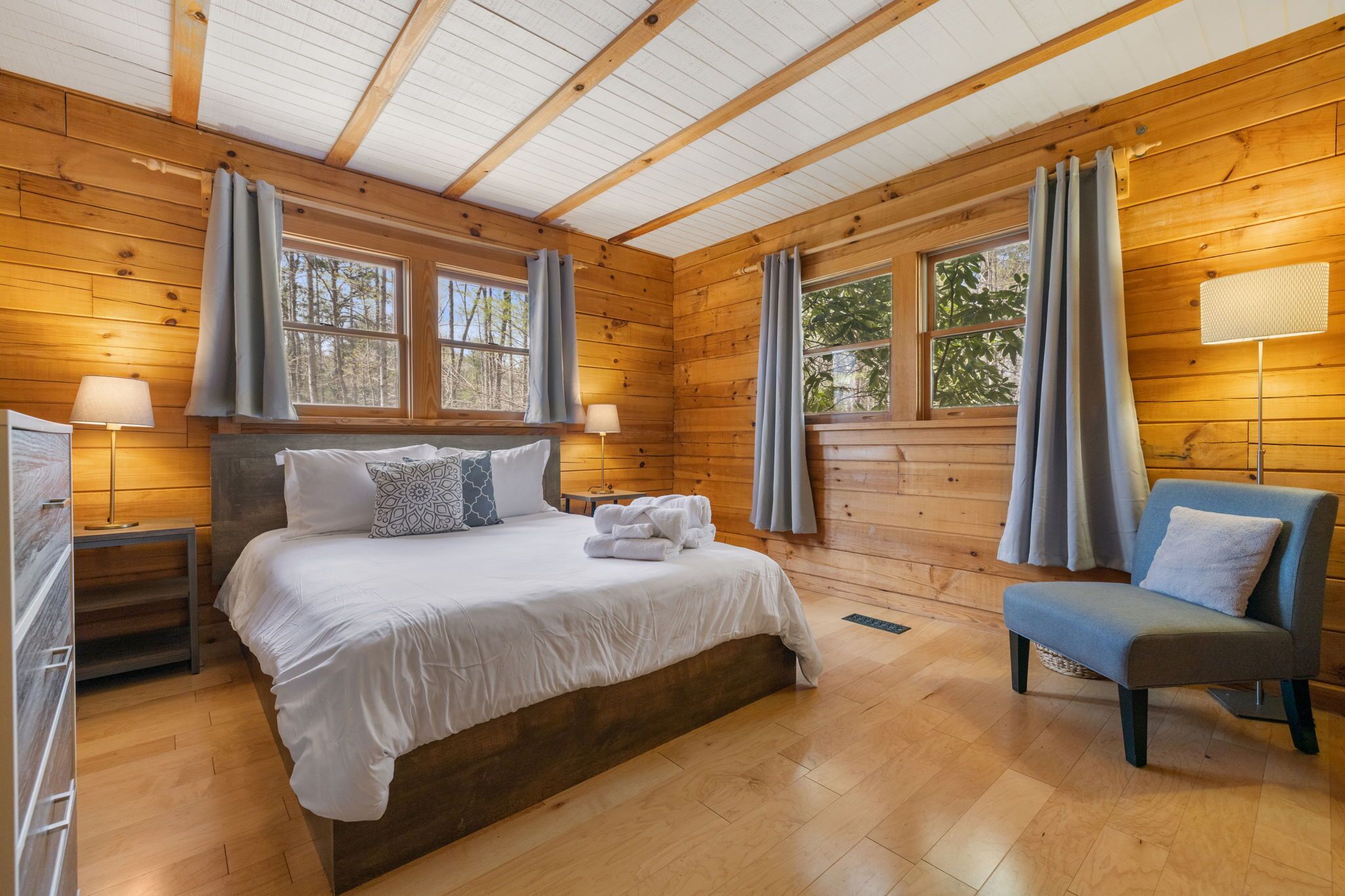 Dormitorio de cabaña de madera con cama, dos mesitas de noche, una silla y ventanas amplias con cortinas.