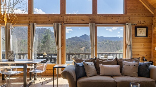 L’intérieur chaleureux d’un chalet, avec un décor en bois, un canapé agrémenté de coussins, une table à manger entourée de chaises et de grandes fenêtres offrant une vue sur les montagnes.