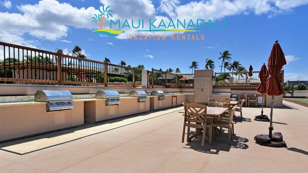 An outdoor barbecue area with multiple grills, wooden tables, and chairs, surrounded by a fence and palm trees. - Top Floor, Corner 2 Bedroom, Ocean View, Beach Cabana, 3 pools, Jacuzzi. (Kaanapali)