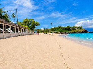 A beach with a building on the left, a clear sky, and footprints leading to the water.