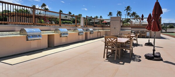 An outdoor barbecue area with multiple grills, wooden tables, and chairs, surrounded by a fence and palm trees.