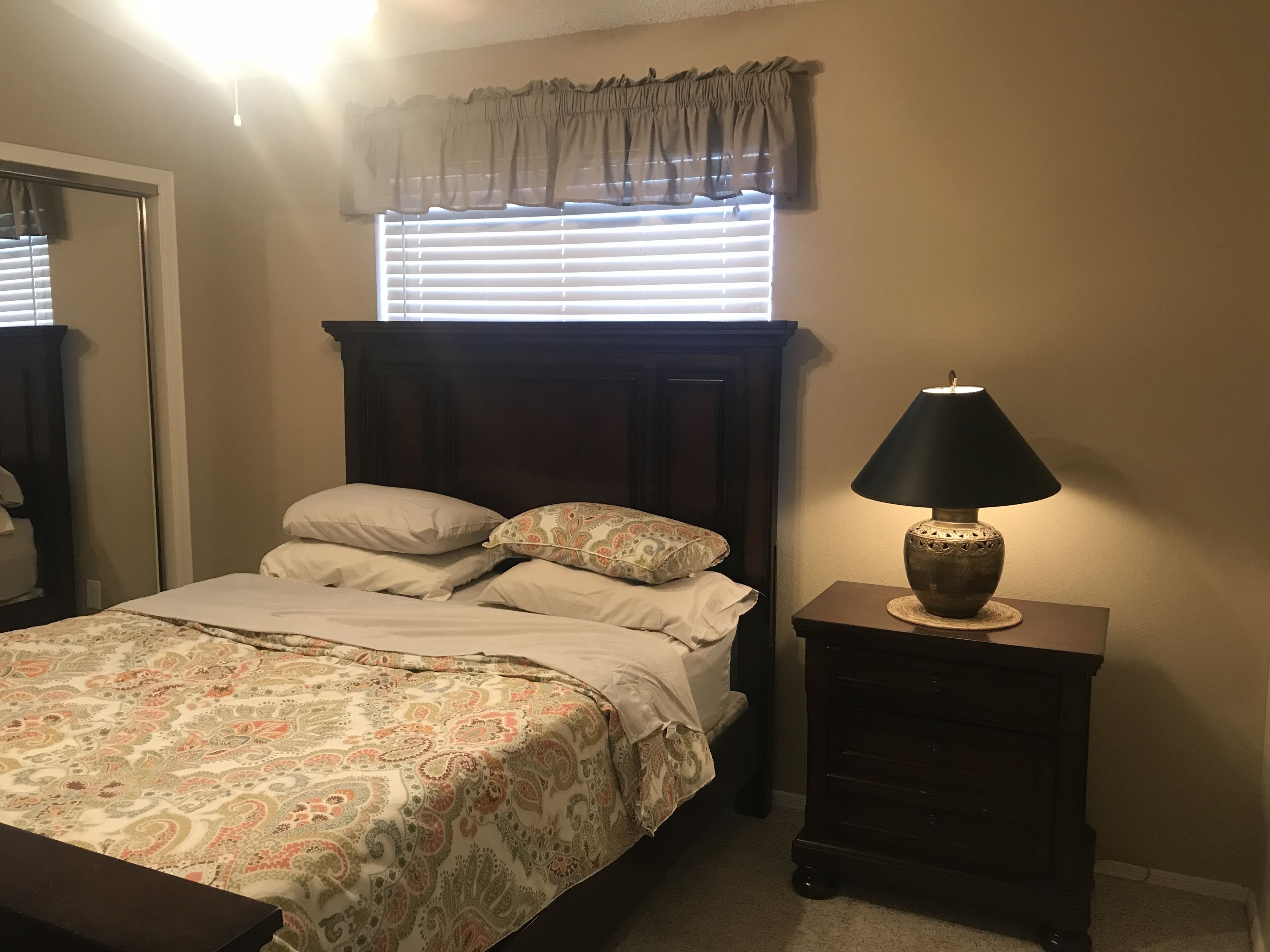 Une chambre à coucher avec un lit, une table de chevet, une lampe, une fenêtre avec des stores et un rideau.