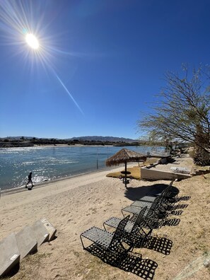 A beach with sun loungers and a thatched umbrella, a clear sky, and a person walking by. - Colorado River Mohave ValleyAZ-Private Bch & Lnch Ramp-Cabral Riverfront Oasis-2 (Mohave Valley)