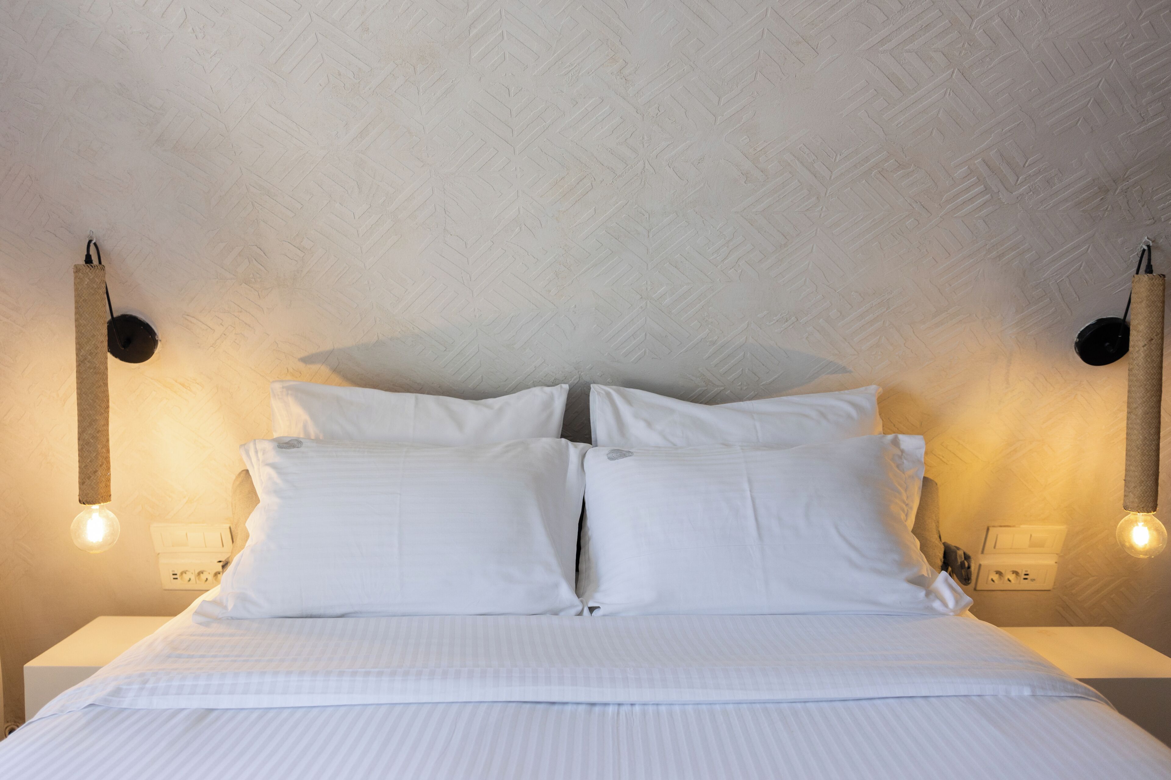 A neatly made bed with white linens, a textured ceiling, and wall-mounted light fixtures.