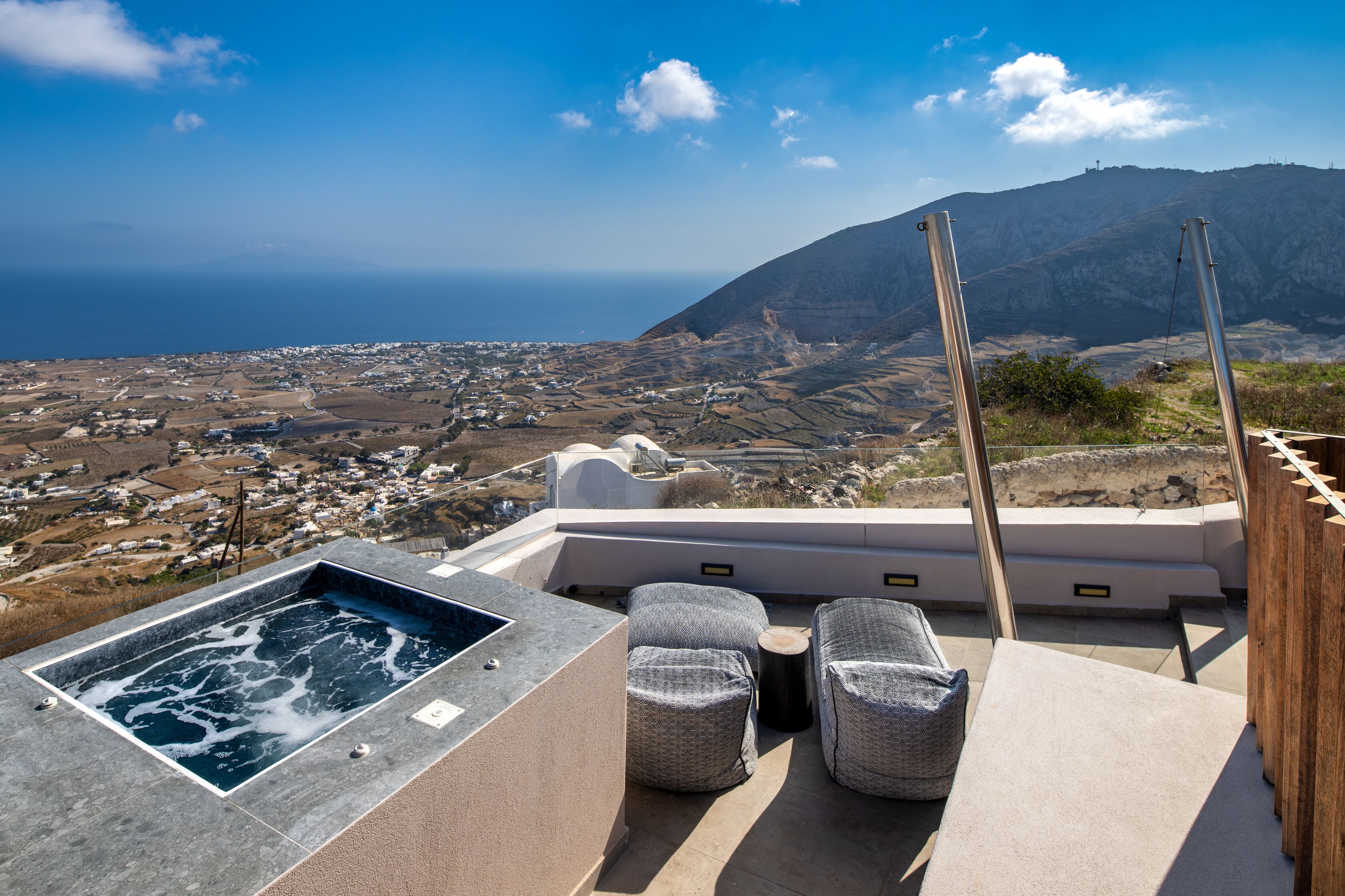 A rooftop terrace with a hot tub, cushioned seating, and a view of a coastal town and mountains.