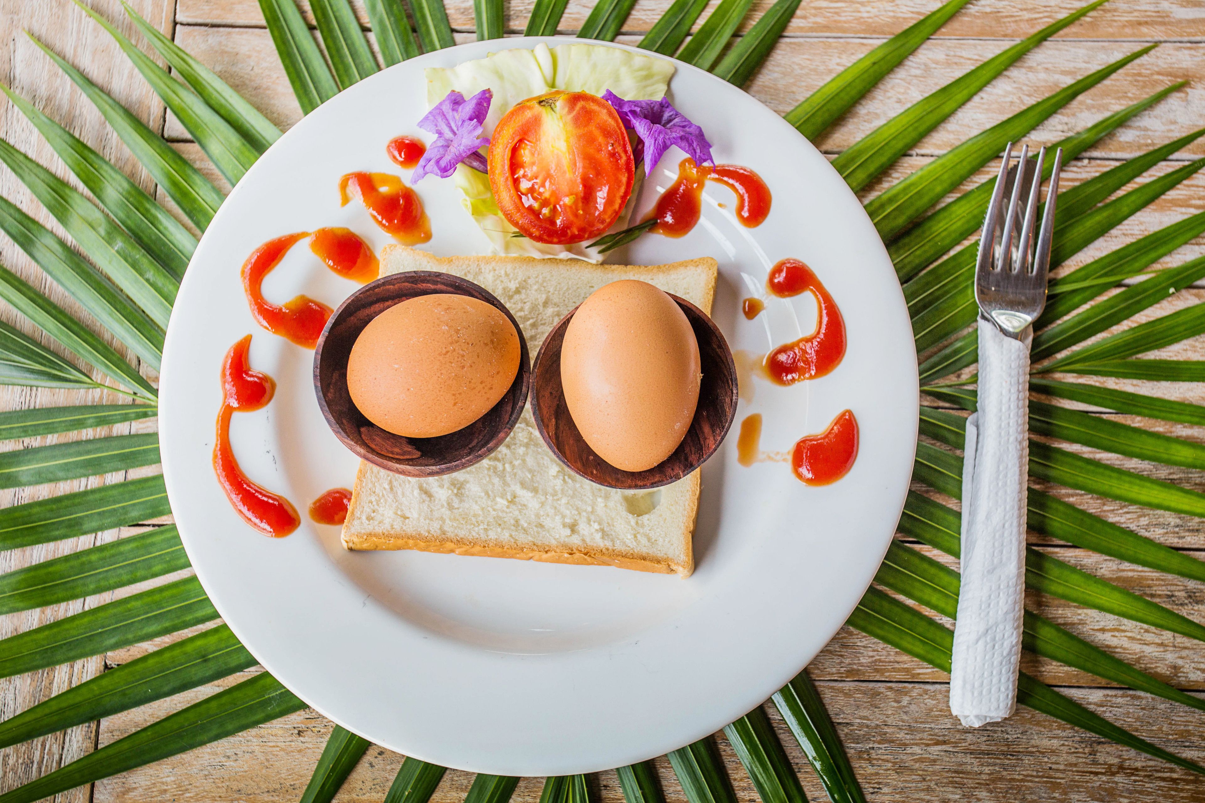 Un plato con dos huevos sobre tostadas, lechuga y rodajas de tomate, rodeado de hojas verdes y un tenedor sobre una mesa de madera.