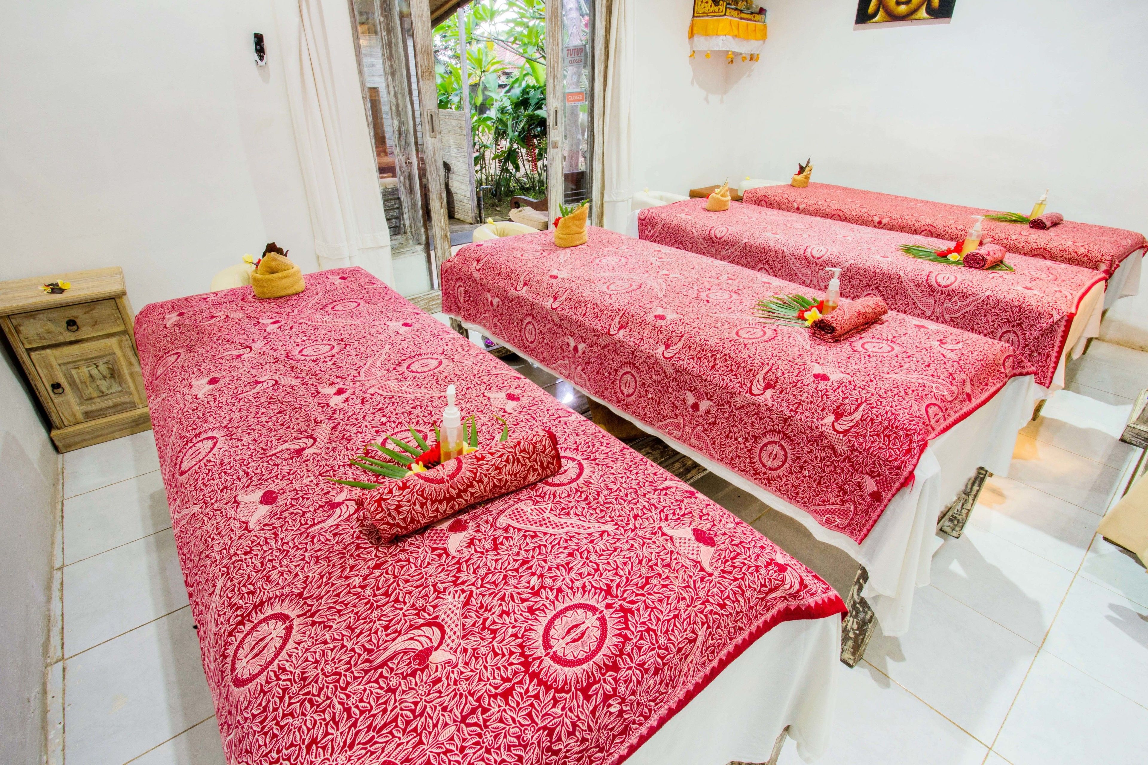 Ein Raum mit drei Massagetischen, die mit rot gemusterten Laken bezogen sind, jeweils mit einem Handtuch und einer Ölflasche.