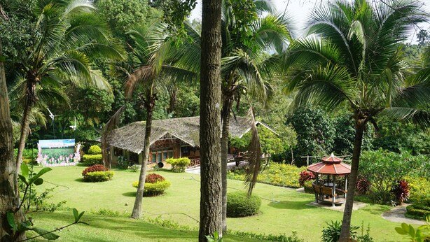 A lush garden with palm trees, a thatched-roof building, and a gazebo. - Rai Aeifoon Resort (Thep Sathit)