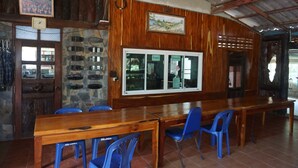 A rustic dining area with wooden tables and blue plastic chairs, stone walls, and a large window with multiple panes. - Rai Aeifoon Resort (Thep Sathit)