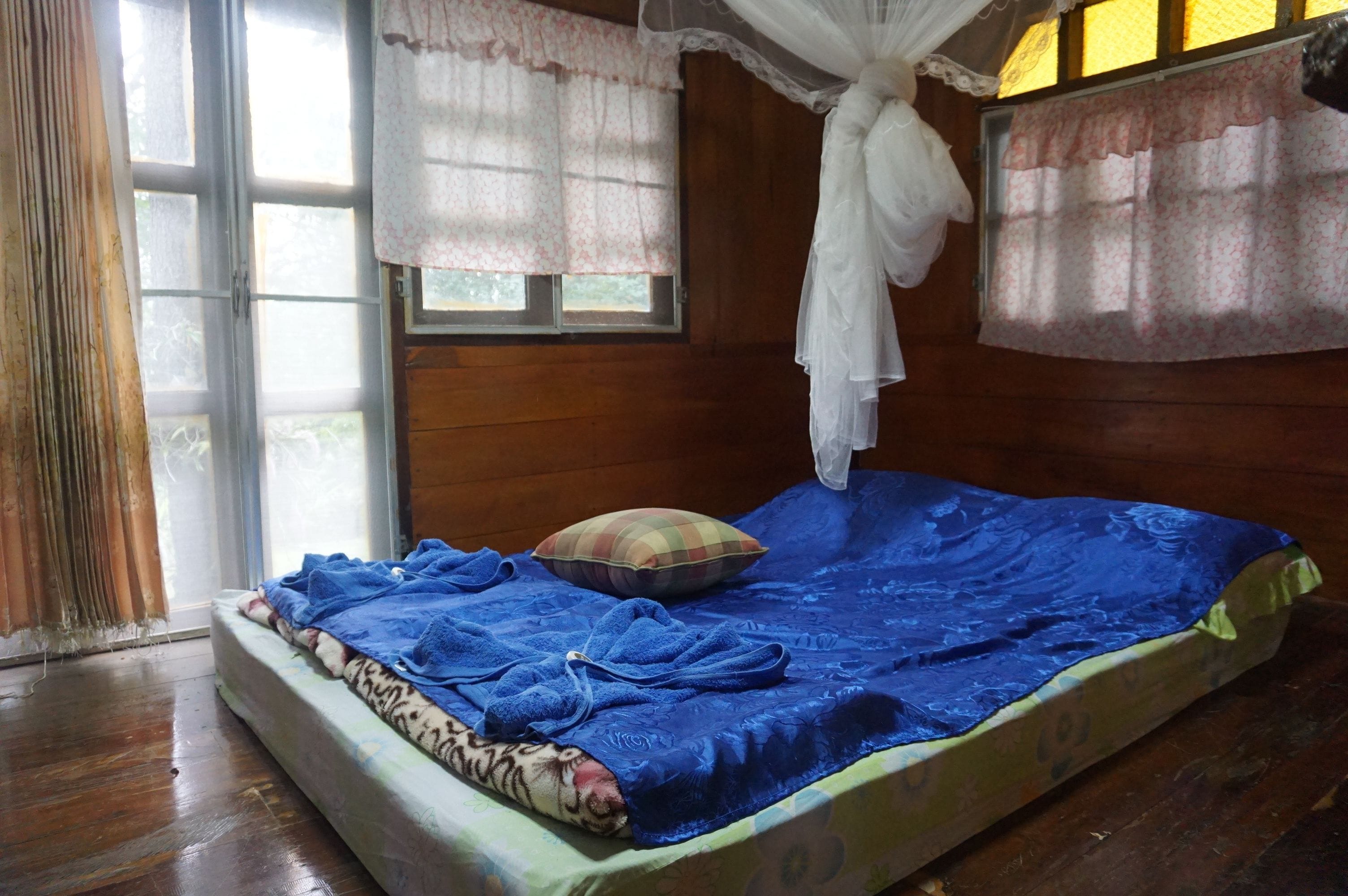 A single bed with a blue blanket and a pillow, a wooden floor, and a window with curtains.