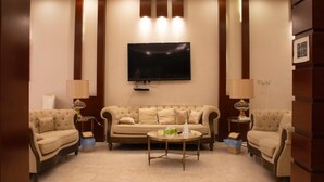 A living room with a large flat-screen TV mounted on the wall, a beige sofa, two armchairs, a coffee table, and two lamps.