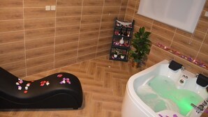 A spa room with a massage chair, a bathtub, and a shelf with bottles and flowers.